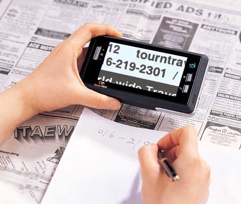 Image of person holding sense view in one hand with the device aimed down at a newspaper. The person is writing down what he sees on the sense view screen with the other hand.